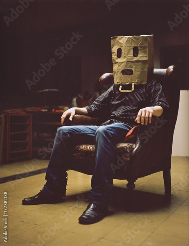 A man sitting on a armchair wearing a spooky paper-bag as a mask.