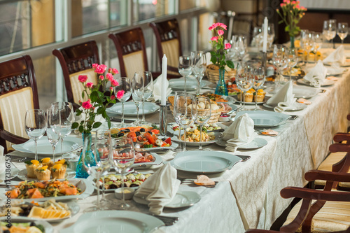 Beautiful festive table served for wedding celebration dinner at home or restaurant interior. Plenty of different food and cutlery. Long table covered with tablecloth and decorated with flowers.