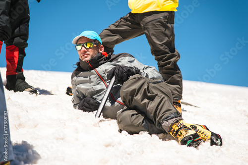 A mountaineer in a sunny spot and professional equipment lying resting on a snowy slope surrounded by his comrades
