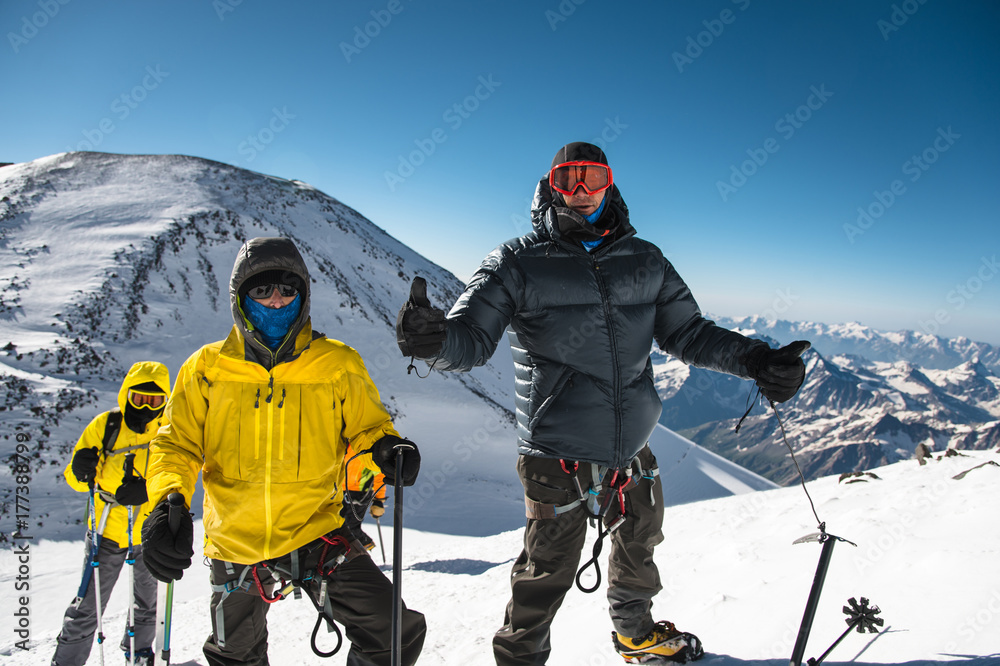 A middle-aged climber in a down jacket and harness shows a thumb up next to his friends on the way to the top of a snow-capped mountain