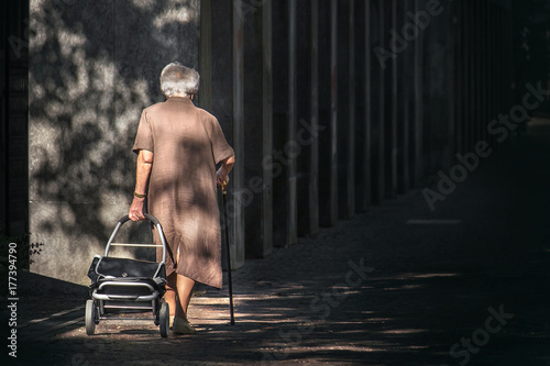 old woman walking alone, senior lady with cane and suitcases with wheels going into the darkness  