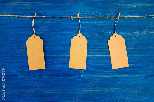 Three brown blank paper price tags or labels set hanging on a rope on the blue wooden background.