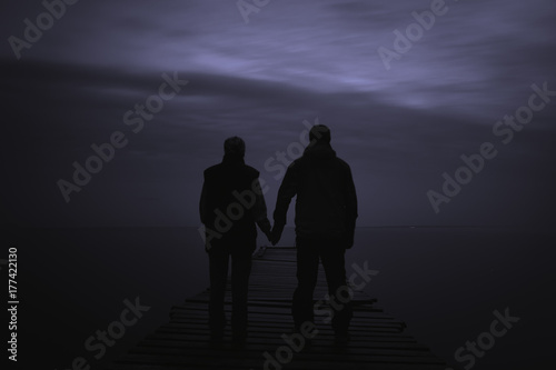 A couple on a sea pier stand at night against a cloudy sky.