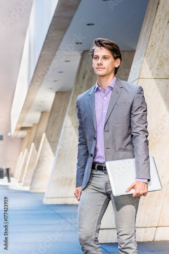 Εκτύπωση καμβά Young Businessman working in New York, wearing gray blazer, patterned shirt, holding laptop computer, walking out from office building to street