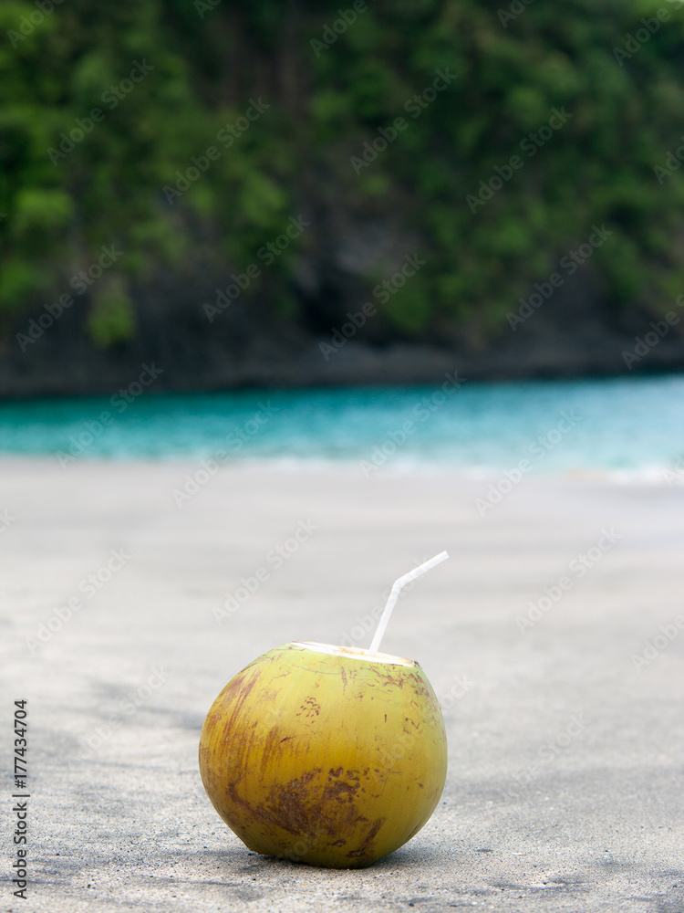 Green coconut with plastic straw on sand beach. Turquoise sea water with white foam and big wave. Powerful and peaceful nature concept. Exotic vacation on the ocean Green coconut with plastic straw on sand beach. Turquoise sea water with white foam and big wave. Powerful and peaceful nature concept. Exotic vacation on the ocean