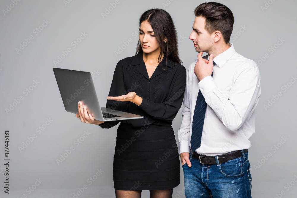 Beautiful business woman and handsome businessman in formal suits are using a laptop, talking and smiling, standing before gray Beautiful business woman and handsome businessman in formal suits are using a laptop, talking and smiling, standing before gray