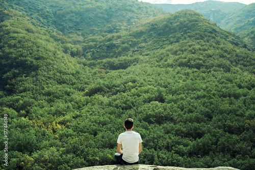 Retrato de hombre joven de espaldas frente a un frondoso verde bosque en la colina de una montaña. El retratado se sitúa en una gran roca observando toda la naturaleza frente a él. photo