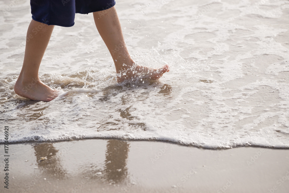 Vacation concept. Close up of boy's  legs walking on the water by the beach. Child by the sea with reflection on wet sand.
