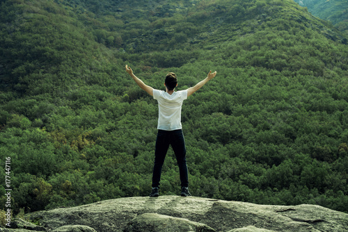 Retrato de hombre joven de espaldas frente a un frondoso verde bosque en la colina de una montaña. El retratado se sitúa en una gran roca observando toda la naturaleza frente a él. photo