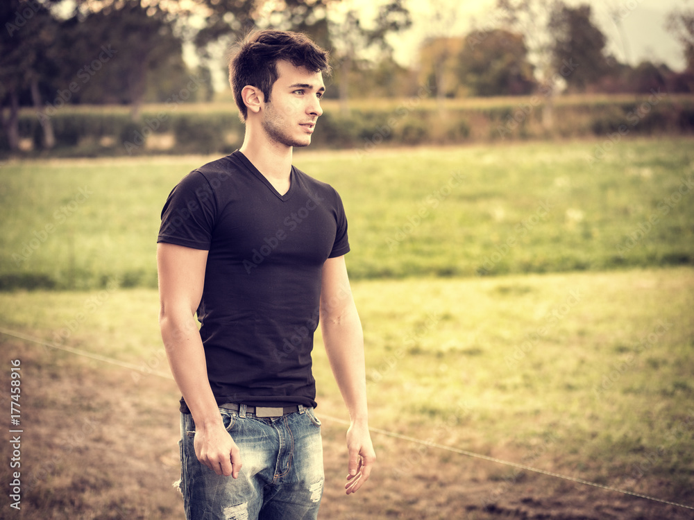 Attractive, fit young man relaxing standing on lawn in the countryside in the grass, looking away Attractive, fit young man relaxing standing on lawn in the countryside in the grass, looking away