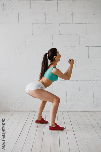 Sports girl doing warm-up, squats. Beautiful girl is training in white studio. Morning exercises. Young woman clothed in aquamarine top, white shorts, red sneackers