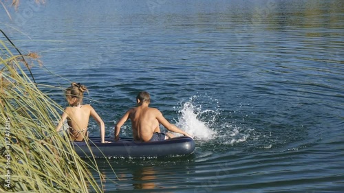 Caucasian teen boy and girl sit on blue inflatable mattress floating on the lake dangling feet in water.