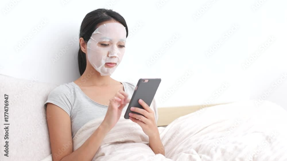 Woman apply facial mask on bed and reading on cellphone Woman apply facial mask on bed and reading on cellphone
