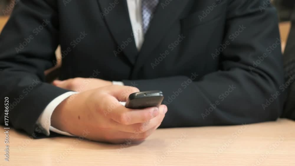 Close-up of unknown young man in black suit sit at desk and holding a black smartphone by pressing his finger on the screen and looks at the important information. Close-up of unknown young man in black suit sit at desk and holding a black smartphone by pressing his finger on the screen and looks at the important information.