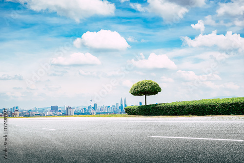 Fototapeta Naklejka Na Ścianę i Meble -  Side view of asphalt road highway with small green trees and grass with modern city skyline in background.
