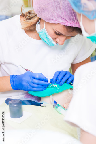 Young beautiful woman dentist in white lab coat and sterile medical cap treats teeth against tooth decay and plaque in child, assistant dentist drops dental instruments in modern dental office