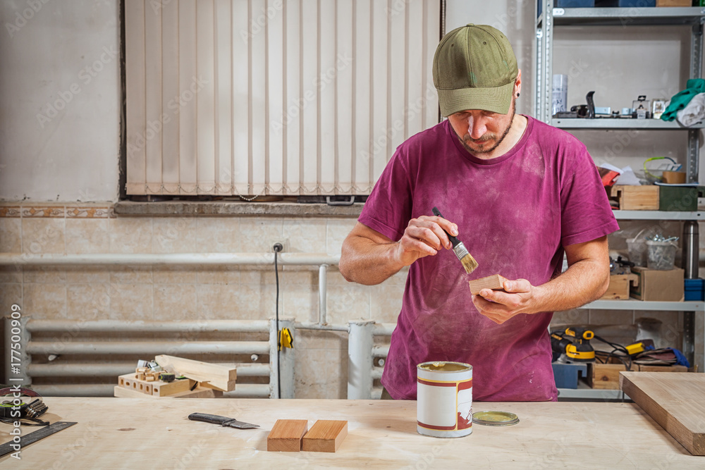 Close-up The man carpenter in working clothes paints a wooden block with a brown protective paint from Corrrosia on a wooden desktop, on which there is a paint, a spatula in the workshop Close-up The man carpenter in working clothes paints a wooden block with a brown protective paint from Corrrosia on a wooden desktop, on which there is a paint, a spatula in the workshop