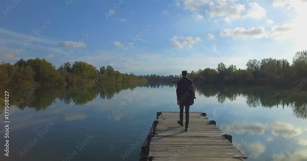 Giovane ragazzo con barba e cappello sta camminando su un molo di legno in riva a un lago Giovane ragazzo con barba e cappello sta camminando su un molo di legno in riva a un lago