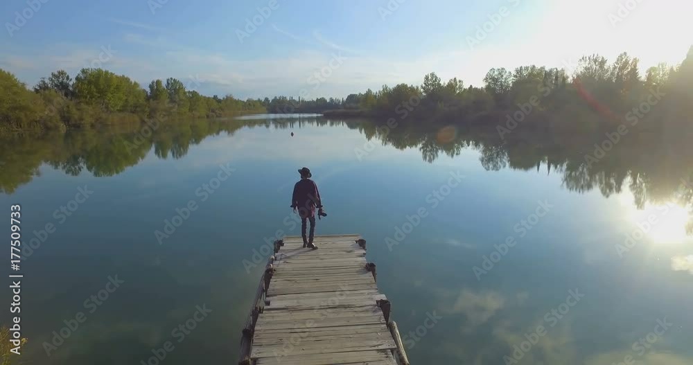 Giovane ragazzo con barba e cappello sta camminando su un molo di legno in riva a un lago Giovane ragazzo con barba e cappello sta camminando su un molo di legno in riva a un lago