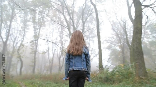 Little European girl with a long hair, blue jacket, black pants, sneakers and blue eyes. A frightened little child is standing in the foggy deserted forest. Loneliness. Steady cam fly around shot.