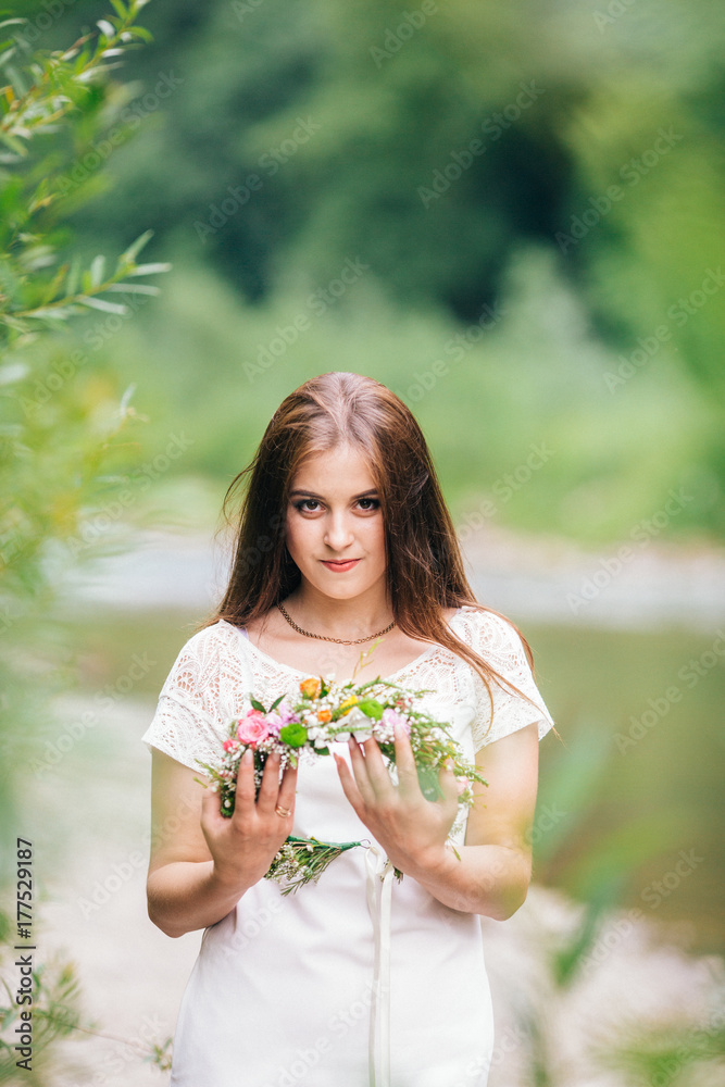 Beautiful young girl in a white chemise and a wreath of flowers on her head sitting stay near mountain river. Beautiful young girl in a white chemise and a wreath of flowers on her head sitting stay near mountain river.