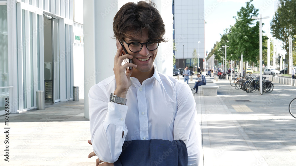 Fototapeta premium Handsome young businessman in city talking on cell phone, also looking at camera, serious