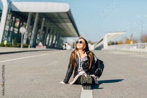 Pretty brunette hipster traveller girl sitting on the middle of asphalt road. Young woman sitting on the high way. Female sitting on center mall of the road. One white line along road.