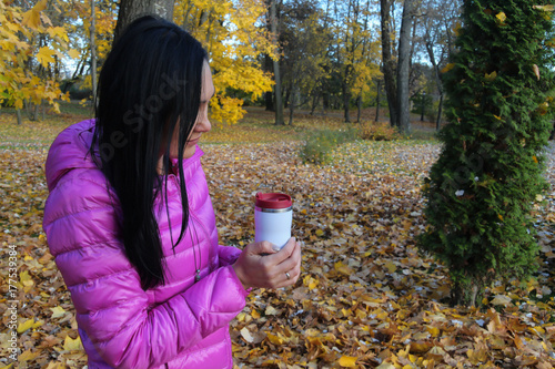woman in bright jacket holds in hands thermo mug in an autumn park