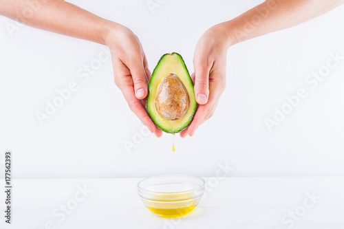 Fresh avocado with oil drop in female hands and bowl of oil on the white wooden background isolated. Healthy, natural beauty concept. Front view. Space for text.
