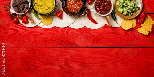 Mexican food concept: tortilla chips, guacamole, salsa, chilli, refried black beans, pulled beef, chicken, cheese and fresh ingredients over vintage red rustic wooden background. Top view

