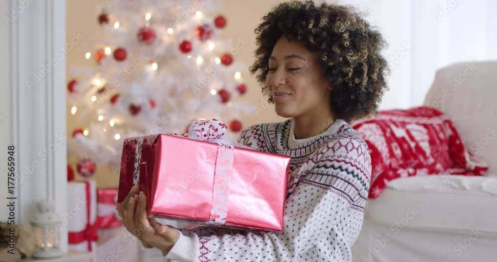 Happy young woman holding a Christmas gift looking down at it with a beaming smile as she relaxes at home in a decorated living room Happy young woman holding a Christmas gift looking down at it with a beaming smile as she relaxes at home in a decorated living room