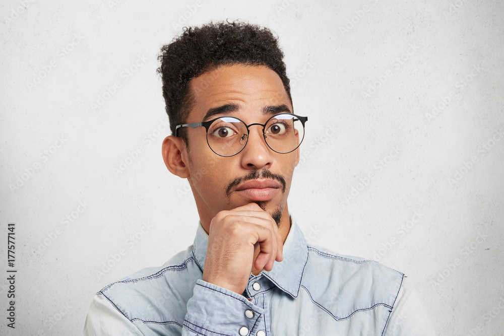 Puzzled confused handsome dark skinned man with Afro hairstyle keeps hand on chin, looks in berwilderment as doesn`t understand something. Attractive businessman wears spectacles and denim shirt