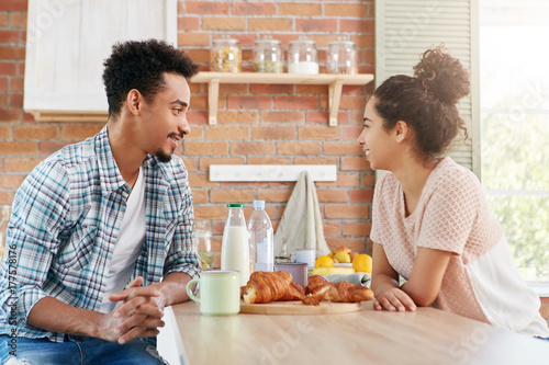 Fotografía Portrait of dark skinned mixed race man visits his female friend, have pleasant conversation, haven`t seen for long time, treats himself with tasty croissant and tea