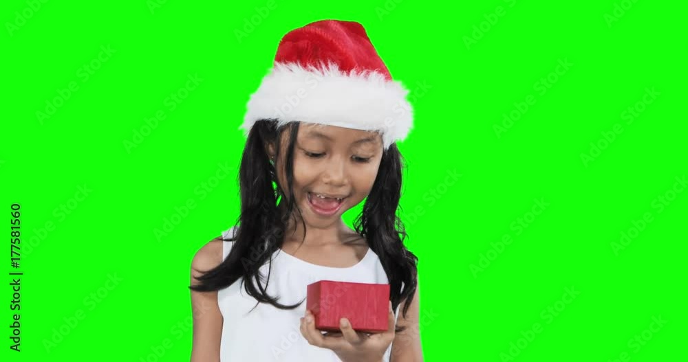 Happy little girl opens a Christmas gift with surprised expression and wearing Santa hat in front of green screen background, shot in 4k resolution Happy little girl opens a Christmas gift with surprised expression and wearing Santa hat in front of green screen background, shot in 4k resolution