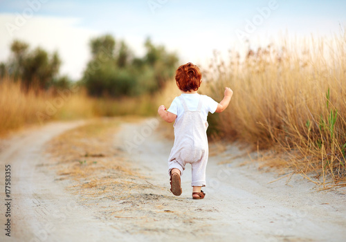 cute toddler baby boy running away along the path at summer field
