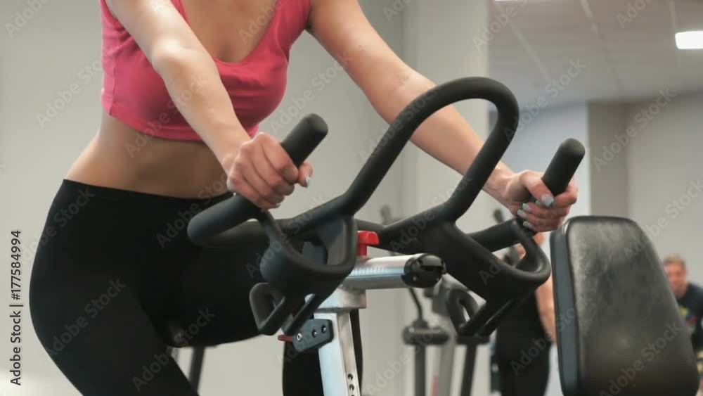 Close-up of unknown of slender blond girl in sportswear is engaged on bike sport simulator in the gym Close-up of unknown of slender blond girl in sportswear is engaged on bike sport simulator in the gym