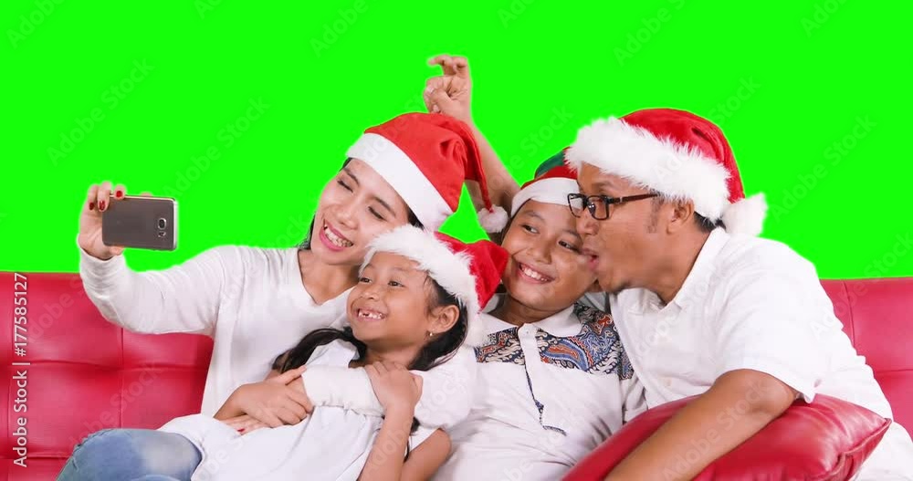 Happy family celebrate Christmas day while wearing Santa hat and taking selfie photo together with a smartphone on sofa, shot in 4k resolution with green screen background Happy family celebrate Christmas day while wearing Santa hat and taking selfie photo together with a smartphone on sofa, shot in 4k resolution with green screen background