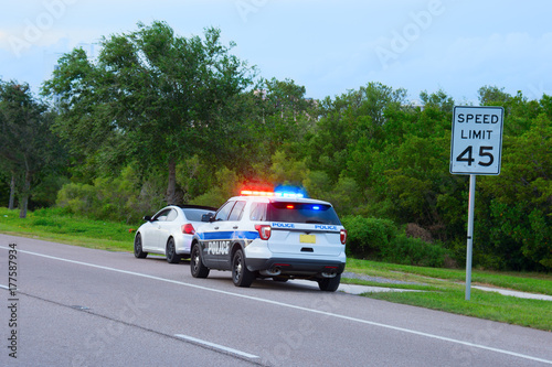 Police truck suv vehicle with flashing red and blue lights has pulled over a sports car for speeding and they happen to be on the side of the road by a speed limit sign.