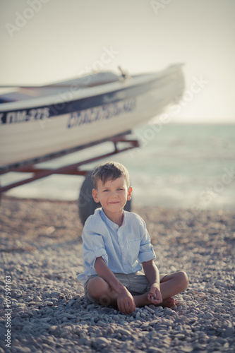 Nice looking handsome boy on beach close to wooden boat wearing fancy stylish blue shirt and shorts with gallows enjoy summer time alone on awesome ocean photo