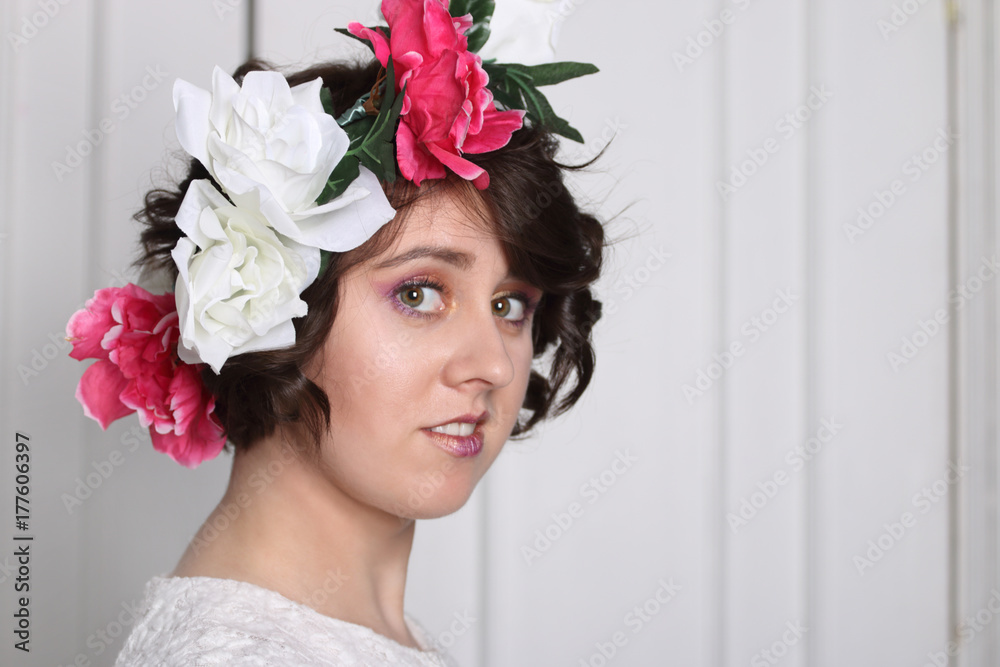 young brunette woman with clean skin, make-up and flower wreath in her hair stands near white door, three quarters