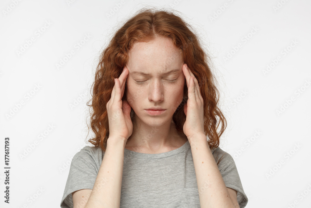 Close up portrait of redhead girl suffering from severe headache, pressing fingers to temples, closing eyes in order to relieve pain with helpless face expression Close up portrait of redhead girl suffering from severe headache, pressing fingers to temples, closing eyes in order to relieve pain with helpless face expression