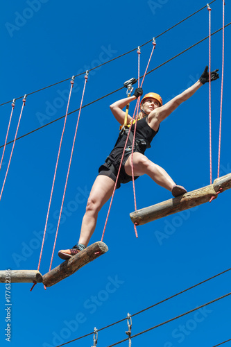 young woman in a helmet with insurance in an adventure climbing park walks by a rope simulator against a blue sky. training mountaineers in the mountains. leisure in nature. sports streets