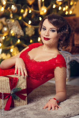 Happy Brunette woman in red dress with gift boxes in luxury interior. Christmas tree. New Year decorations.