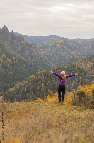 A girl in a lilac jacket jumps on a mountain, a view of the mountains and an autumnal forest overcast. Free space for text