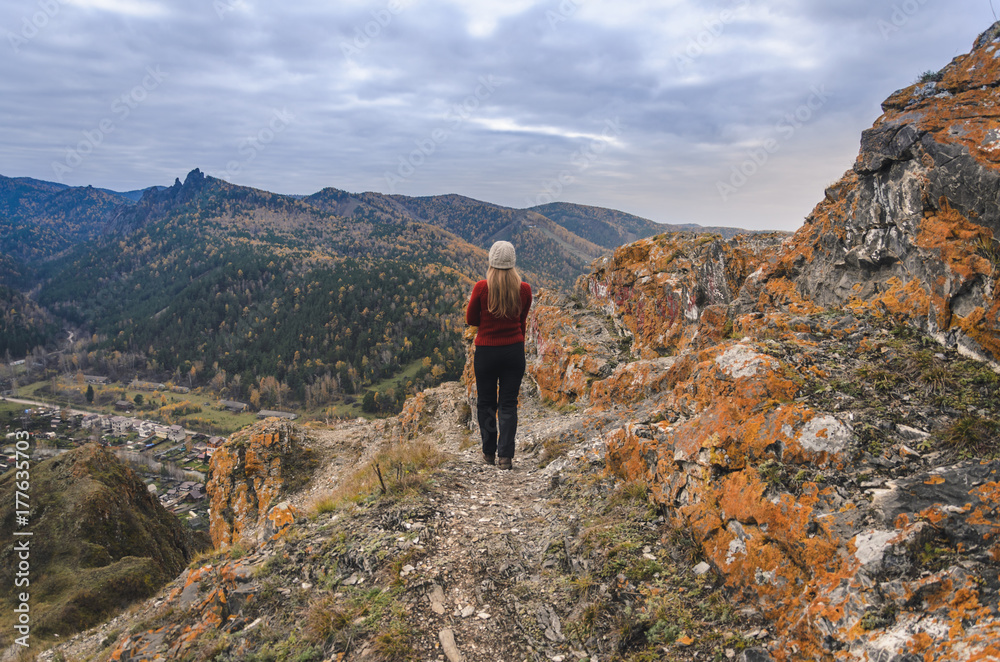 A girl in a red jacket looks out into the distance on a mountain, a view of the mountains and an autumnal forest by an overcast day. Free space for text