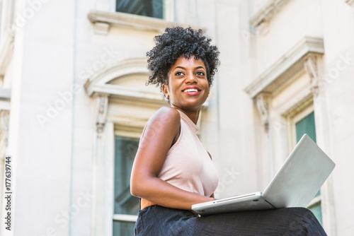 Way to Success. Young African American woman with afro hairstyle wearing sleeveless light color top, sitting by vintage office building in New York, working on laptop computer, looking up, smiling.. photo