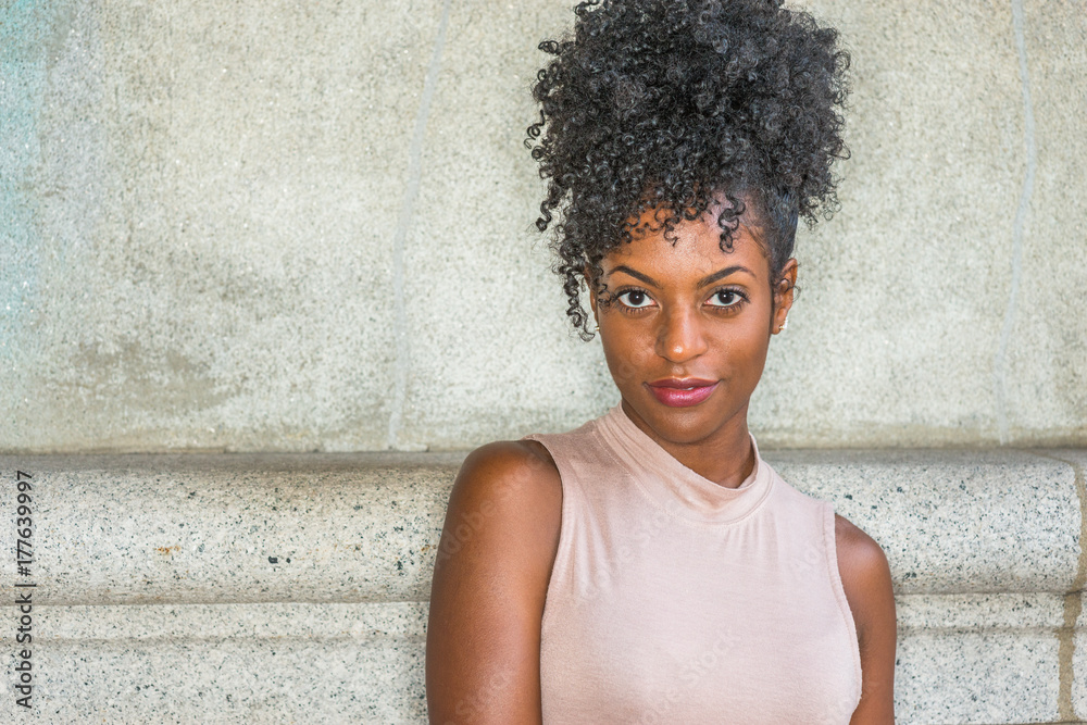 Portrait of Young African American Woman in New York. Young black female college student with afro hairstyle wearing sleeveless light color top, standing against vintage wall, looking at you..