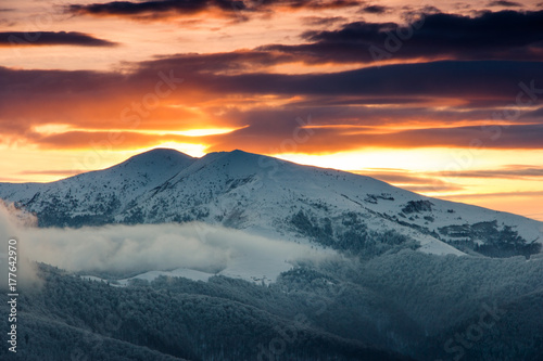 Beautiful sunrise in the winter mountains. Dramatic cloudy over sky. View of foggy hills and trees covered with rime. 