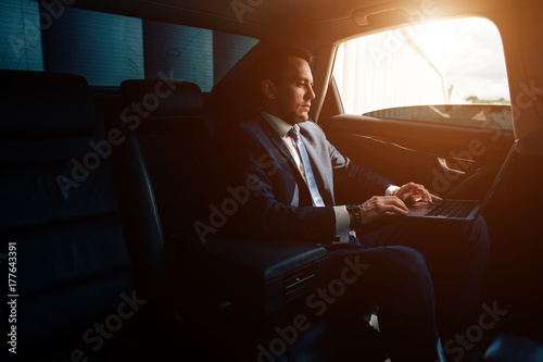 Handsome businessman talking with phone sitting with laptop on backseat of car photo