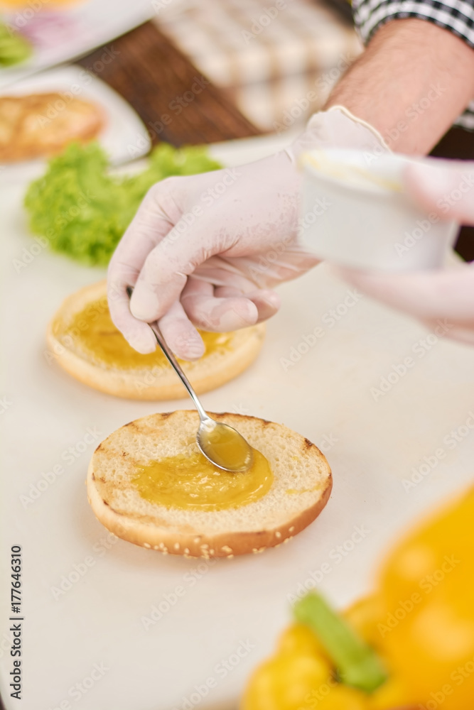 Chef hand spreading the mustard on bun for burger. Cook preparing hamburger at kitchen. Culinary and cooking concept.